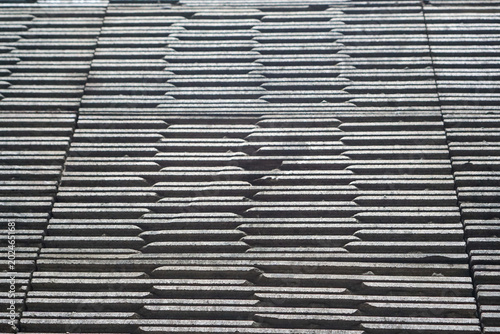 Rows of dark, textured roof tiles stretch across the frame. Each tile shows subtle wear and weathered edges. Uniform pattern creates a rhythmic, industrial visual texture. Ideal for architectural