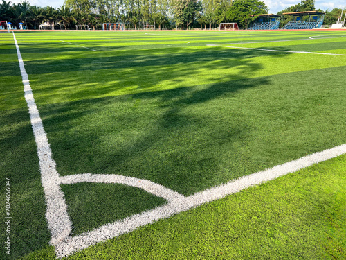 Wide angle view of green artificial grass soccer field with white corner line under a sunlight. Authentic outdoor stadium with stands and trees for sports competition and training backgrounds.