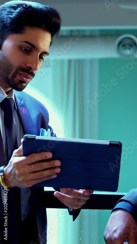 Indian businessmen using tablet computer near glass railing in modern office, Asian young corporate executives collaborating on digital strategy, sharing ideas and analyzing data together