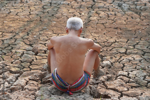 Tableau sur toile A Man in Drought-Stricken Land: An elderly man with white hair sits with his back to the camera, his arms crossed, on cracked, dry earth