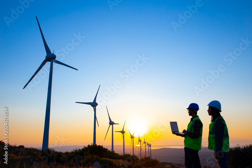 Fotografie The engineers, review data on a laptop while standing near a row of wind turbines on a grassy hill