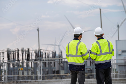 Two professional engineers analyzing renewable energy production at a wind turbine farm power plant substation. High-quality technical inspection and maintenance concept for sustainable electricity.
