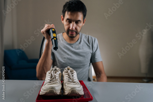 Tableau sur toile Portrait of young bearded man steam cleaning dirty white sneakers on red tray, restoring bright, fresh appearance while demonstrating careful at home shoe care and stain removal routine