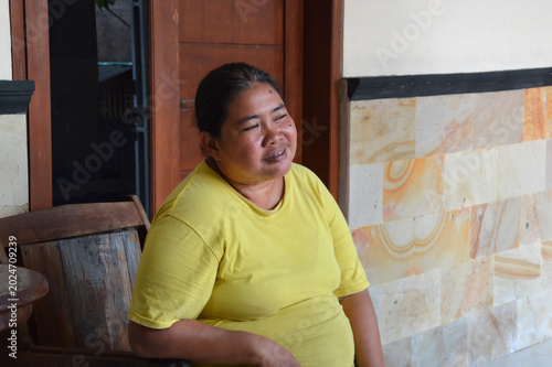 A pleasant middle aged Asian woman wears a yellow shirt while sitting relaxed and smiling gently on a wooden chair on her house veranda.