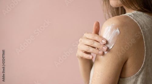 Mujer joven aplicando crema hidratante en el hombro. Concepto de cuidado de la piel, dermatología y belleza, sobre fondo rosa pastel con espacio para texto.