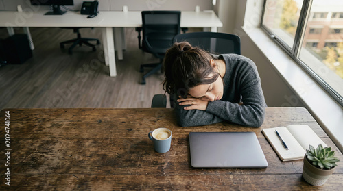 Exhausted woman rests head on hands at wooden desk with laptop, coffee, notebook, and succulent plant near window in modern office space.