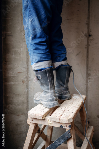 Konstfotografi Close-up of a builder's dusty black work boots standing on a makeshift wooden ladder near a red brick wall during interior construction