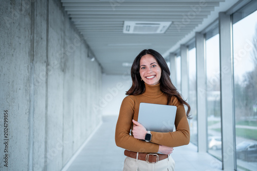 Smiling businesswoman holding laptop walking in modern corridor