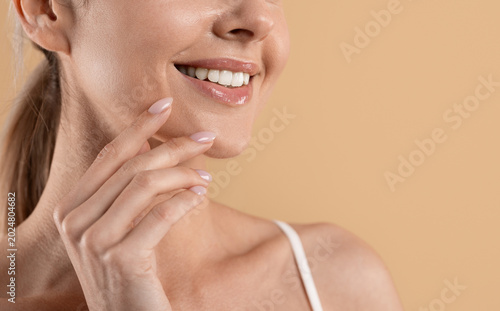 Φωτογραφία Smiling young woman touching smooth glowing skin with hand while standing isolated on beige studio background
