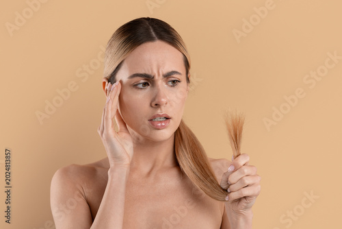 Worried young woman looking at damaged hair ends while checking hair condition, concerned female standing on beige studio background. Beauty and haircare concept
