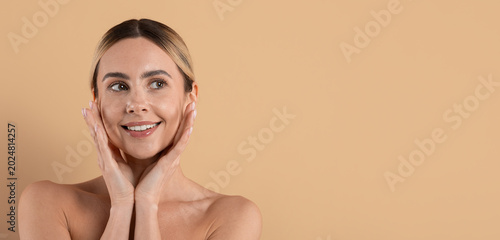 Beautiful young woman touching her face with healthy glowing skin and looking aside, natural skincare and beauty portrait isolated on beige studio background with copy space