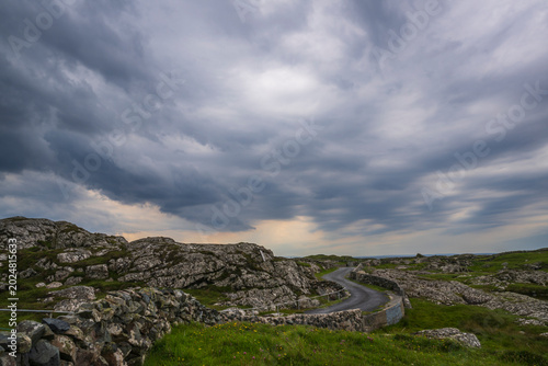 Winding country road through rugged rocky terrain on Lettermullan Island, Connemara, County Galway, Ireland. Rural single track lane under a dramatic storm sky.