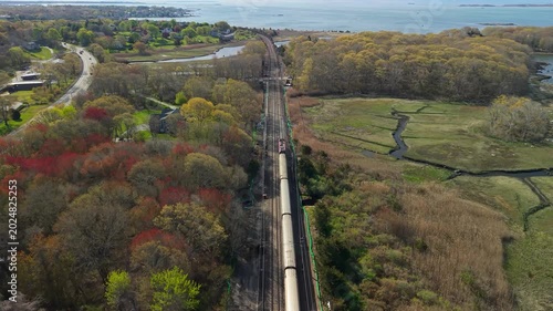 Amtrak passenger train in Mystic aerial view, a high-angle drone tracking shot following a regional train as it travels along the coastal Northeast Corridor tracks in Mystic, Connecticut.