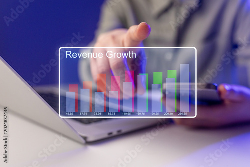 Person analyzing revenue growth data on a laptop while using a smartphone in an indoor setting at night