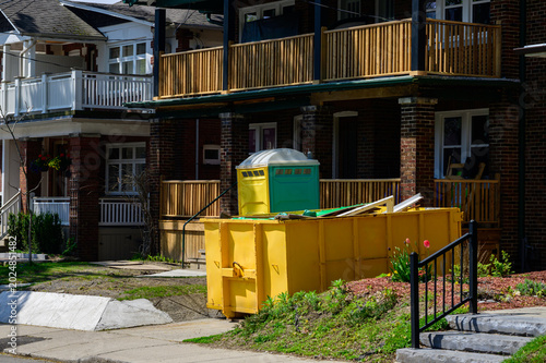 Roll-off dumpster and portable toilet outside a residential gut renovation project on a brick rowhouse street, Toronto, spring.