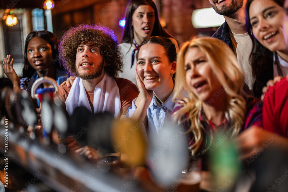 Fototapeta premium Cheerful young fans looking at a screen and celebrating a soccer match victory in a pub.