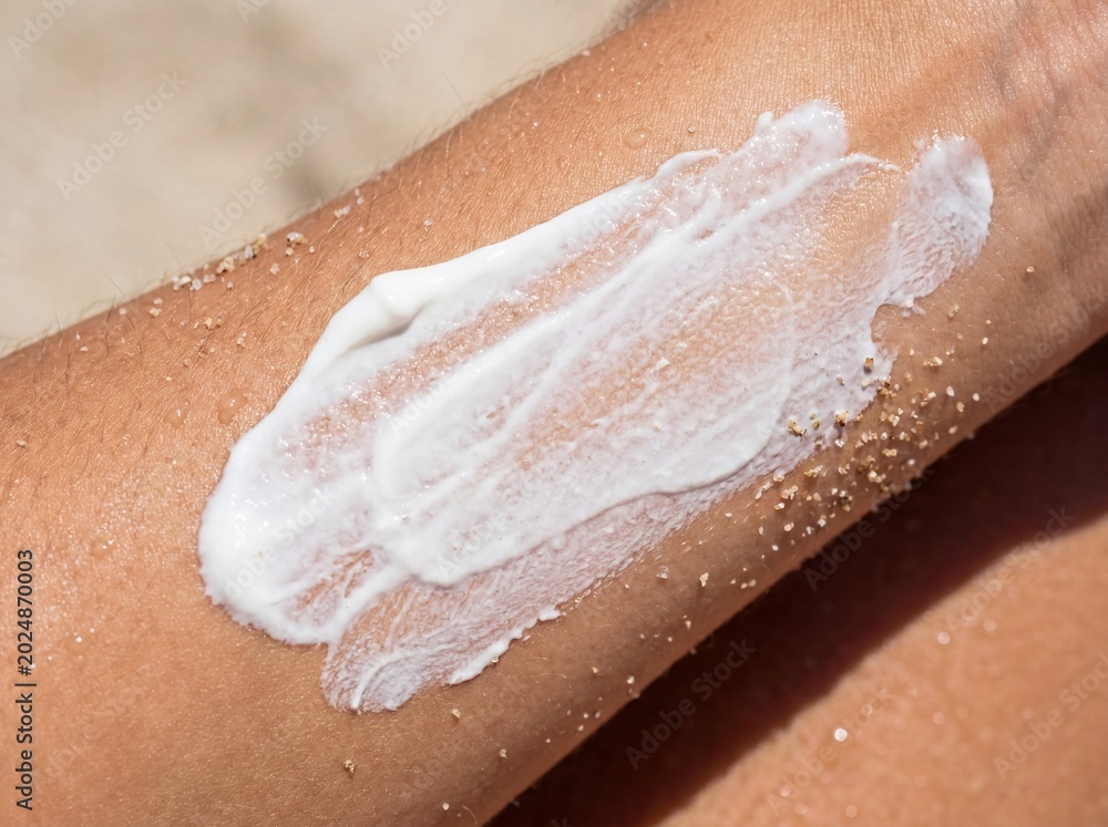 Fototapeta premium Primer plano de crema protectora solar mineral de zinc aplicándose en un brazo bronceado con arena y cristales de sal marina.