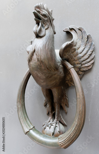 Striking close-up of an ornate cast bronze door knocker shaped as a proud Gallic rooster the Coq Gaulois standing atop a ring handle, beak open mid-crow.