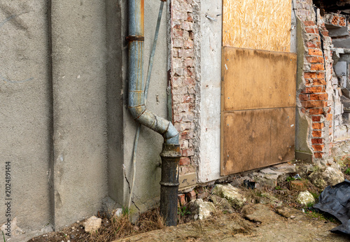 Old metal drainpipe attached to a cracked plaster and exposed brick wall beside a boarded doorway and crumbling masonry, highlighting architectural decay, abandoned building detail, and urban deterior