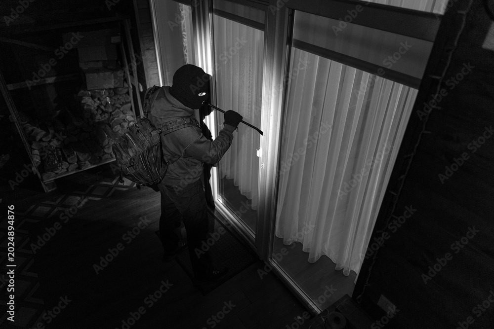 Fototapeta premium Man burglar in dark clothing trying to unlock door or window of house using crowbar