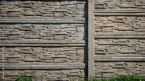 Close up view of a beige precast concrete fence panel featuring a stacked stone texture. Vertical posts separate sections. Green grass grows along the bottom in sunlight