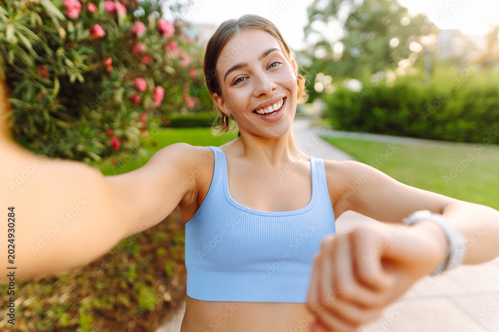 Fototapeta premium Portrait of a smiling woman in sportswear taking a selfie in a sunny park. A young athlete enjoys relaxing after a workout. Concept for sports and blogging. Active lifestyle.