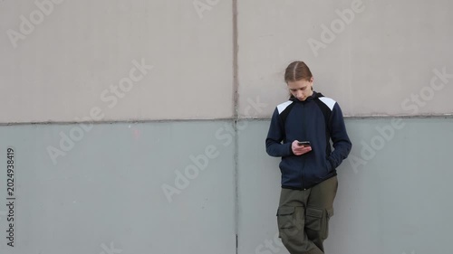 Urban teen with phone. Young person leaning against wall. Casual adolescent standing near cityscape background. Youthful person in informal posture resting by urban stone structure