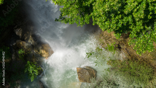 Photos A dramatic aerial view of a majestic waterfall cascading into a crystal-clear turquoise river, nestled between a dense forest and a rocky canyon