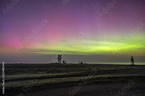 The historic white foghorn tower and the red-and-white Winkie Lighthouse stand under a vast night sky painted with green and pink Northern Lights,Point of Ayre, Isle of Man.