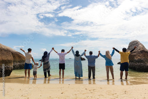 Multi-generational family standing together on a tropical beach with raised hands facing the ocean, capturing happiness, unity, vacation moments, and strong family bonding under a bright blue sky.