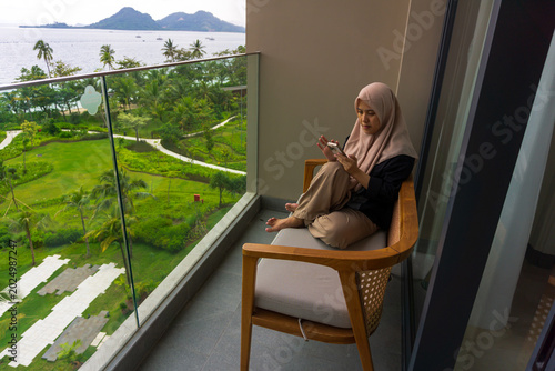 A beautiful Asian woman wearing a hijab sits on a hotel balcony chair while holding a cell phone. A depiction of a strong, thoughtful, and caring woman on vacation with her family.