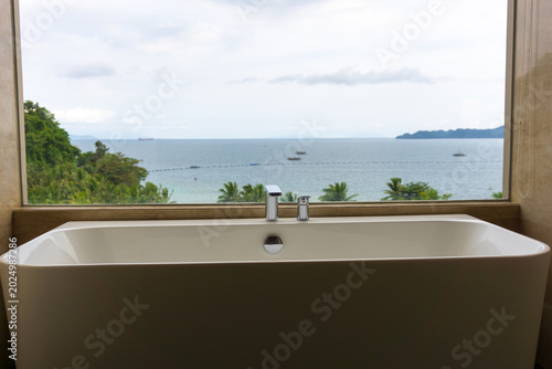 Modern freestanding bathtub with a panoramic ocean view through a large window, creating a luxurious and relaxing tropical resort atmosphere with serene coastal scenery and minimalist interior design.