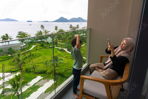 Portrait of a mother sitting on a hotel balcony chair holding a cell phone while taking a photo of her son with a beautiful view. A happy and warm atmosphere during a family vacation.
