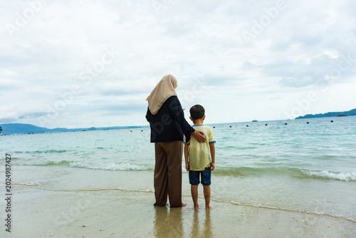 Portrait of a mother and son standing on the beach looking at the calm sea while chatting with their child, a calm and comfortable atmosphere during the holidays.