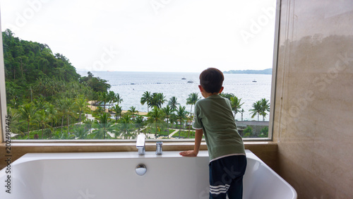 Photo of a boy standing in a bathtub looking at the view of the sea and beautiful islands from the bathroom window of a hotel in Indonesia.