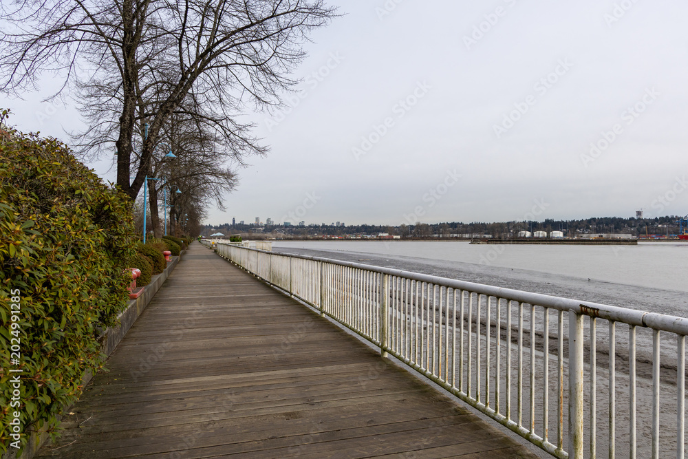 Fototapeta premium Riverside Boardwalk Promenade With Bare Trees And Distant City Skyline In New Westminster