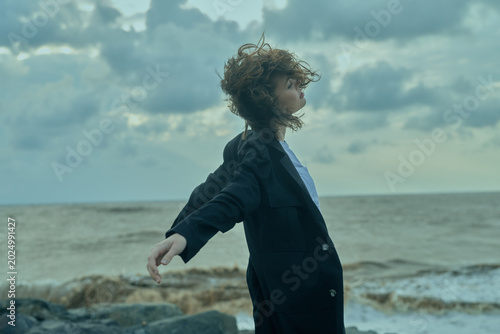 Schilderij op canvas Business, wind, sea, jacket scene with a figure standing on a rocky shore, coat fluttering, open posture and expansive horizon, empowering mood