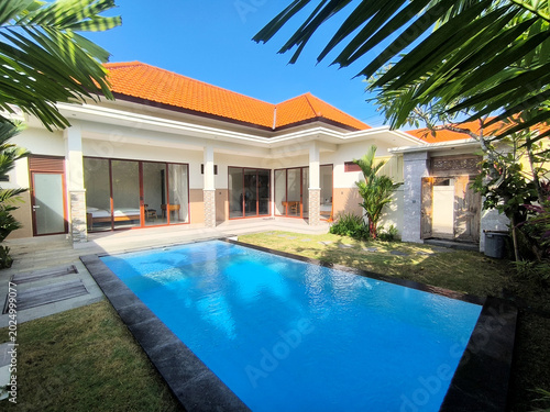 Canvas-taulu Beautiful private pool in a modern Balinese villa with orange tile roof, surrounded by lush tropical gardens and traditional carved wooden doors, under bright blue skies
