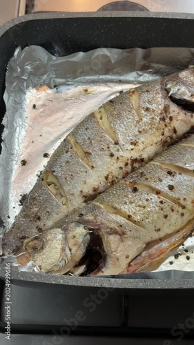 Roasted bass black pan on stove, close-up of two seasoned sea fish resting on foil after oven cooking, speckled pepper crust, warm