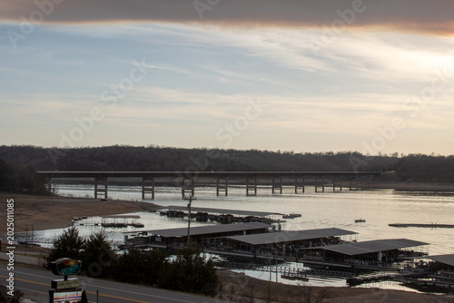 Tablou pe pânză a wide scenic overlook of a concrete girder highway bridge stretching across a wide river, featuring a boat marina with covered docks in the foreground under a soft sunrise or sunset sky