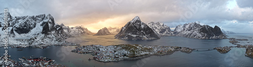Aerial panoramic view of snow-covered mountains and scenic fjords in northern Norway. Golden-hour light glows through the distant clouds, adding warmth and drama to the Arctic landscape and calm water