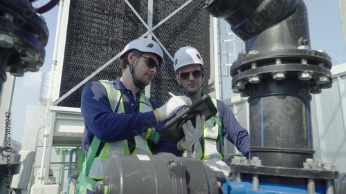 Engineer maintenance hvac cooling tower inspection rooftop industrial plant with technician team checking pipe, valve, and pump via tablet checklist, safety vest and helmet, secure service daylight
