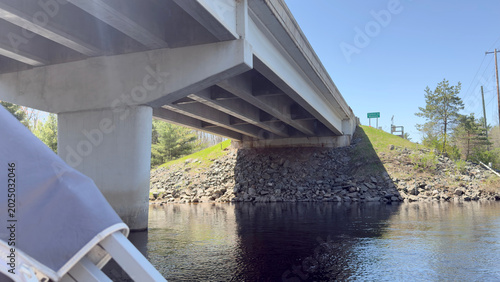 Quadro su tela low-angle perspective taken from a watercraft, looking upward at the structural underside of a modern concrete girder highway bridge crossing over a river channel under a bright sky
