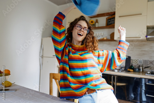Photos Cheerful woman in a colorful knit sweater dances in a bright kitchen, throwing a