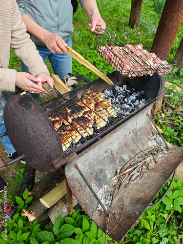 Two individuals preparing and cooking tasty marinated meat, including chicken pieces and raw pork or beef, on a vintage, weathered barrel-style barbecue grill with burning coals in a vibrant natural