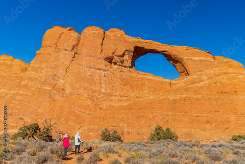 Fotografie Hikers admire a natural sandstone arch against a blue sky.