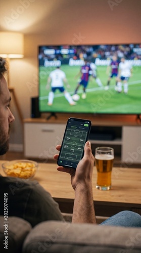 Close-up of a hand using a sports betting and statistics mobile app on a smartphone while watching a live soccer match on TV