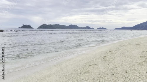 Serene tropical beach with gentle waves and distant islands under a cloudy sky.