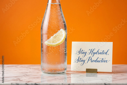 Glass bottle of sparkling water with a lemon slice next to a motivational card on a marble table © Meow Creations