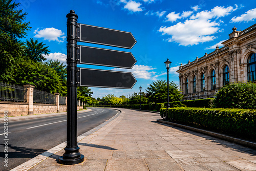 Blank black directional signpost with three arrows on a city street. Empty street sign template with copy space for text. Classic architecture building and blue sky background